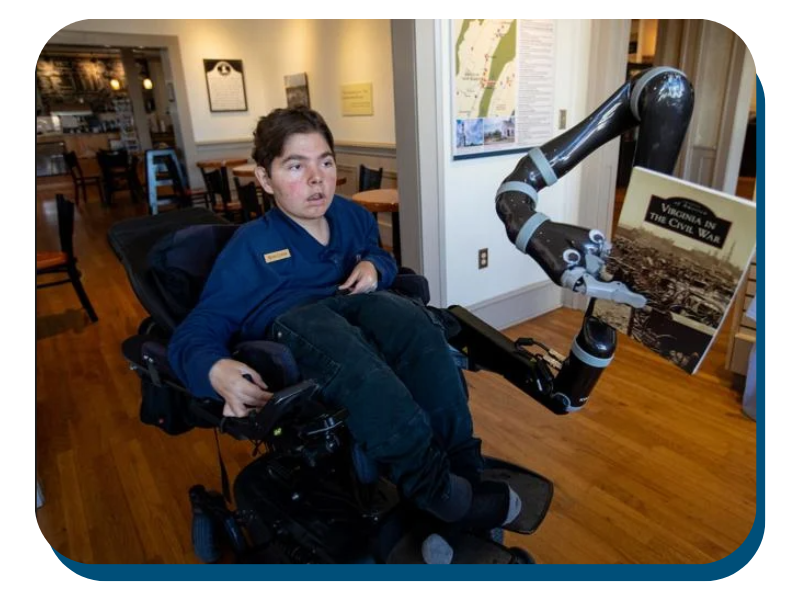Wade Custer uses a new robotic arm on his wheelchair to pick up a book in the New Market Battlefield Visitor Center showing his new robotic arm
